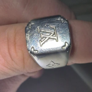 Louis Vuitton silverring med LV-logga - Snygg signetring från Louis Vuitton i silverfärg med LV-logga på ovansidan och ikoniska blommor i hörnen. Ringen har en fyrkantig form och är tillverkad i siiver med en polerad finish. Perfekt statement-piece för dig som gillar lyxiga accessoarer.
