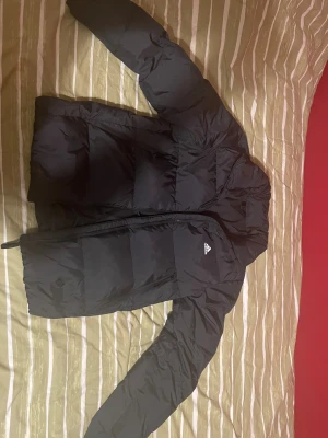 Svart pufferjacka från Adidas - Svart Adidas pufferjacka i nyskick. Använd endast ett par gånger och köpt för någon månad sedan inför vintern, men blev aldrig använd för vintern då jag köpte en ny jacka. Kvitto finns tyvärr inte kvar, men modellen kostar ca 80 £ (runt 1000 kr) ny.  Mitt pris: 200 kr – kan gå ner vid snabb affär. Jackan är helt fräsch utan fläckar eller skador. Fler bilder kan skickas vid behov.  Hör gärna av dig om du har frågor