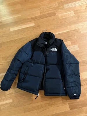 Svart pufferjacka från The North Face - Säljer en mörkblå pufferjacka från The North Face med hög krage och logga på bröstet. Jackan har dragkedja framtill och två sidofickor med dragkedja. Perfekt för kalla dagar och har en stilren, sportig look.