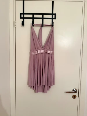 Ljuslila kort klänning från Vero Moda - Supersöt ljuslila kort klänning från Vero Moda med djup v-ringning, knyt i nacken och ett glansigt band med rosett i midjan. Kjolen är plisserad och har ett flowigt fall. Perfekt för dig som vill sticka ut med en feminin och trendig look.