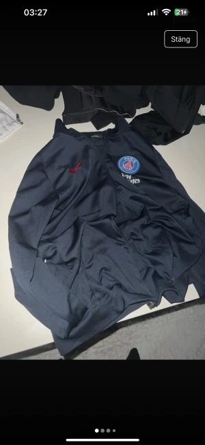 PSG Nike Dri-Fit träningsjacka L - Säljer en mörkblå Paris Saint-Germain träningsjacka från Nike med Dri-Fit-teknologi. Jackan har röd Nike-logga, PSG-emblem på bröstet, Fly Emirates-tryck och en röd detalj längs sidan. Materialet är syntet och jackan har dragkedja framtill.