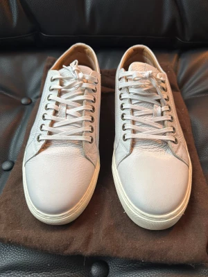 Vita sneakers från Oscar Jacobson - Fräscha vita sneakers från Oscar Jacobson med rund tå och platt sula. Skorna är tillverkade i mjukt skinn med snygg struktur och har klassisk snörning. Insidan är beige och sulan är vit med svart undersida. Perfekta för dig som gillar stilrena och tidlösa sneakers.