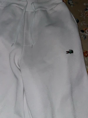 Vita mjukisbyxor från Lacoste - Snygga vita mjukisbyxor från Lacoste med klassisk krokodillogo broderad på benet. Byxorna har snörning i midjan och är tillverkade i mjukt bomullsmaterial. Passformen är normal och benen är smalare nedtill för en modern look.