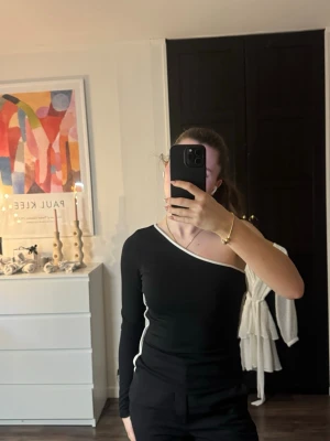Svart oneshoulder topp med vit kant - Snygg svart oneshoulder topp med lång ärm och vit kant längs halsringningen och sidan. Toppens passform är tight och den har en stilren, modern look som passar perfekt till svarta byxor eller jeans. Materialet känns mjukt och stretchigt mot kroppen.