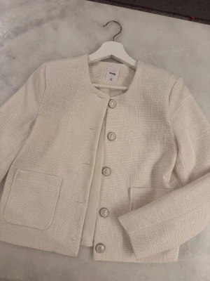 Beige kavaj från Sinsay XS - Stilren beige kavaj från Sinsay i storlek XS. Jackan har rund halsringning, dekorativa silvriga knappar framtill och två fickor. Materialet är strukturerat och ger en exklusiv känsla. Perfekt för dig som gillar en klassisk och chic look.