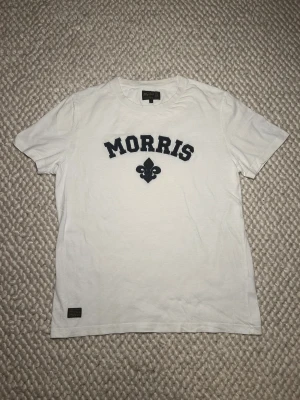 Vit t-shirt från Morris med tryck - Vit t-shirt från Morris med klassiskt svart tryck på bröstet och en broderad fleur-de-lis. T-shirten har rund hals, korta ärmar och är gjord i mjuk bomull. Perfekt för en clean och stilren look.