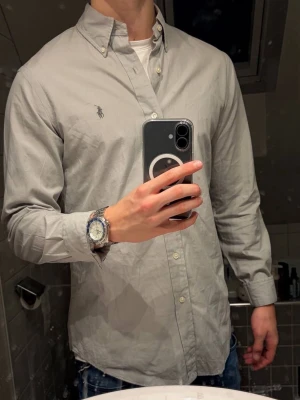  Skjorta från Polo Ralph Lauren - Snygg grå skjorta från Polo Ralph Lauren med klassisk button-down krage och broderad logga på bröstet. Custom fit-modell i mjuk bomull, perfekt för dig som gillar stilren och enkel design. Långärmad med vita knappar framtill. Skjortan är i väldigt bra skick, använd få gånger. Ny pris 1699 kr. Priset kan absolut diskuteras!