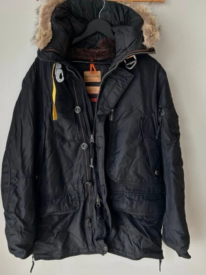 Svart Parajumpers vinterjacka med päls - Svart Parajumpers vinterjacka med huva och avtagbar päls, flera fickor och snygga detaljer som gul rem och patch på ärmen. Jackan har dragkedja, knappar och fodrad insida för extra värme. Perfekt för kalla dagar och riktigt stilren look.