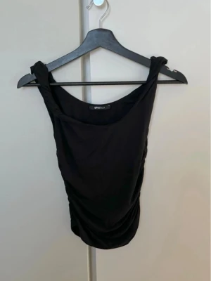 Svart linnetopp med knyt från Gina Tricot - Svart linnetopp från Gina Tricot i storlek S. Toppen har breda axelband med snygg knyt-detalj och drapering längs ena sidan för en cool look. Materialet är mjukt och känns skönt mot huden. Perfekt att styla med jeans eller kjol för en trendig vibe.
