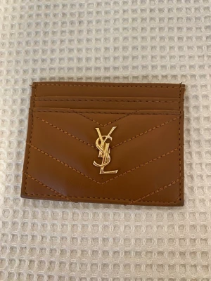 Brun YSL plånbok i skinn - Snygg brun plånbok från YSL i äkta skinn med guldigt YSL-emblem framtill. Plånboken har flera kortfack och stilrena sömmar i chevronmönster. Perfekt för dig som vill ha en lyxig och stilren accessoar i fickformat.