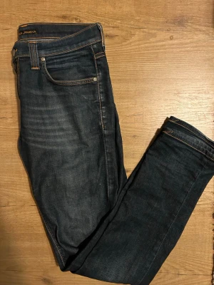 Mörkblå jeans från Nudie Jeans - Snygga mörkblå jeans från Nudie Jeans med klassisk femficksdesign och kontrastsömmar i orange. Jeansen har rak passform och är tillverkade i slitstarkt denimtyg. Perfekta för dig som gillar stilrena och tidlösa jeans.