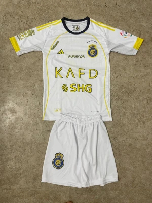 Al Nassr Ronaldo fotbollskit barn - Snyggt Al Nassr fotbollskit för barn med Ronaldo 7-tryck på ryggen. Setet är vitt med gula detaljer och klubbmärke på både tröja och shorts. Tillverkat i lätt polyester för bästa komfort på planen. Perfekt för unga fotbollsfans som vill spela som sin idol.