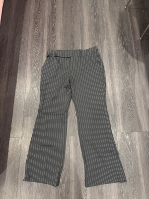 Svarta randiga kostymbyxor med knappar - Snygga svarta kostymbyxor med tunna vita ränder och två knappar i midjan. Byxorna har raka ben och klassisk passform, perfekta för en stilren look. Materialet känns mjukt och följsamt, och midjan är normalhög. Passar dig som gillar en clean och tidlös vibe.