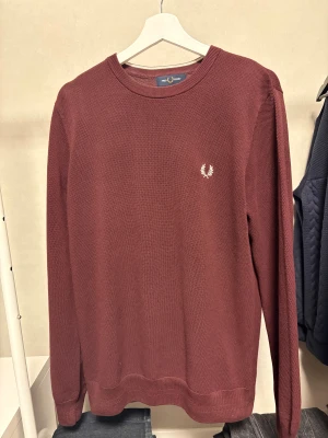 Vinröd tröja från Fred Perry - Snygg vinröd tröja från Fred Perry med klassisk rund hals och diskret logga broderad på bröstet. Tröjan är stickad i ett mjukt material och har långa ärmar samt ribbade muddar vid hals, ärmslut och nederkant. Perfekt för dig som gillar stilrena plagg. Helt ny endast testad en gång!