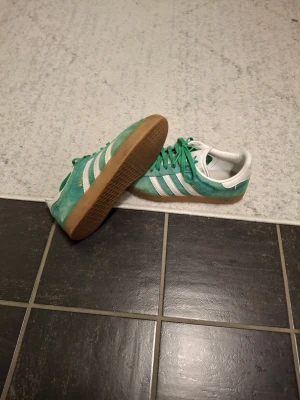 Adidas Gazelle gröna sneakers - Säljer ett par gröna Adidas Gazelle sneakers med klassiska vita ränder och gummisula. Skorna har snörning och är tillverkade i mocka med vit detalj vid hälen. Perfekt för dig som gillar retrostil och vill sticka ut med färg.