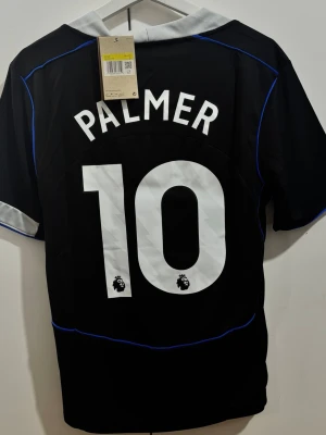 Chelsea tredjeställ 25/26 #Palmer 10 - Chelsea tredjeställ Palmer 10 fotbollströja . svart med vita och blå detaljer. Snyggt broderat klubbmärke på bröstet, Premier League-tryck och Palmer 10 på ryggen. V-ringad krage i grått och korta ärmar. Perfekt för dig som älskar fotboll och Chelsea. TVEKA INTE ATT SKICKA EN BUD