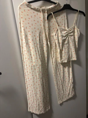 Krämfärgade mjukisbyxor med persikomönster - Säljer ett par krämfärgade mjukisbyxor med sött persikomönster i orange och grönt. Byxorna har snörning i midjan och är tillverkade i mjukt bomullsmaterial. Perfekt för chill dagar hemma eller som mysplagg.