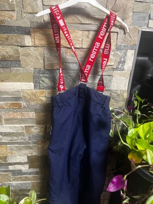 Mörkblå täckbyxor från Reima, strl 134 - Mörkblå täckbyxor från Reima med röda hängslen med logga. Byxorna har elastisk midja, knapp och dragkedja framtill samt slitstarkt yttertyg. Perfekta för kalla dagar och vinteraktiviteter.