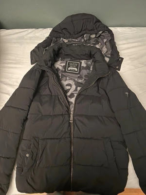 Svart pufferjacka från Guess - Svart pufferjacka från Guess med avtagbar huva och coolt camouflagemönstrat foder i grå toner. Jackan har dragkedja framtill, två fickor med knapp och är tillverkad i syntetmaterial som är både vattenavvisande och vindtät. Perfekt för kalla dagar.