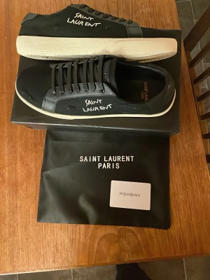 Svarta Saint Laurent sneakers - Svarta sneakers från Saint Laurent med vit handskriven logga på sidan. Skorna har rund tå, platt vit sula och snörning. Materialet är en mix av skinn och textil, vilket ger en lyxig och stilren vibe. Kommer med originalpåse och kort.