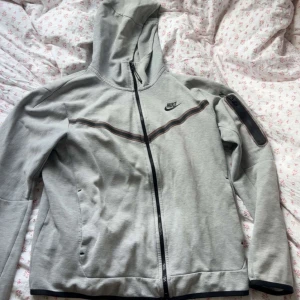 Grå Nike tech - Grå Nike tech  med hel dragkedja, huva och svart nike logga på bröstet. Koftan har en svart detalj längs bröstet och en ficka med dragkedja på ärmen. 