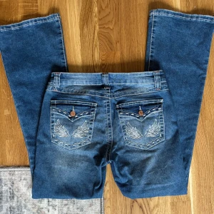 Blå bootcut jeans med broderade fickor - Coola blå bootcut jeans med coola broderade vingar på på bakfickorna med knappstängning. Jeansen har en lätt tvättad look och är perfekta för dig som gillar detaljer.