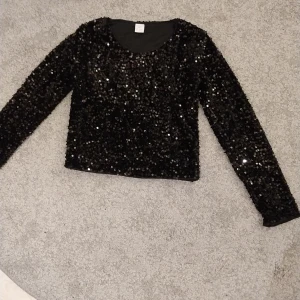 Svart paljettopp från Lindex - Glittrig svart topp från Lindex med lång ärm och rund hals. Hela toppen är täckt av svarta paljetter som ger en riktigt festlig vibe. Materialet är mjukt och stretchigt, perfekt för att sticka ut på nästa event. Passformen är normal och toppen är croppad i modellen.