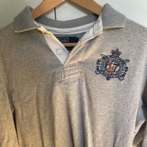 Grå pikétröja från Polo Ralph Lauren - Snygg grå långärmad pikétröja från Polo Ralph Lauren med klassisk krage, knappar och broderad logga på bröstet. Tröjan har en stilren look och är gjord i mjuk bomull, och passar till allt.  Perfekt för dig som gillar Ralph Lauren och vill se bra ut. Säljer då den inte passar mig. Pris diskuterbart, så kom med förslag. 