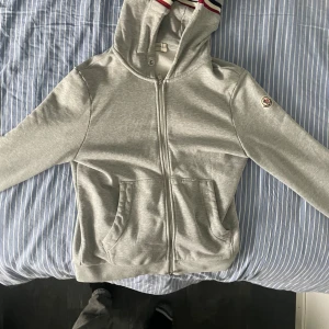 Grå Moncler hoodie med dragkedja - Snygg grå hoodie från Moncler med hel dragkedja och huva. Tröjan har Moncler-logga på ärmen och fickor framtill. Insidan är mjuk och huvan har coola ränder i rött, blått och vitt. Perfekt för chill dagar och streetstyle.