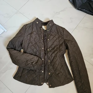 Quiltad brun jacka från lft, M - Quiltad brun jacka från lft med ståkrage och guldfärgade tryckknappar. Jackan har dragkedja framtill, ribbade detaljer vid sidorna och ärmarna samt ett rutigt mönster över hela ytan. Perfekt för dig som gillar en klassisk men ändå chill stil.