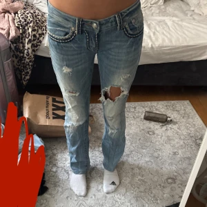 Bootcut jeans med slitningar, stl 28 - Säljer ett par ljusblå bootcut jeans från Miss Me med coola slitningar och hål på knäna. Jeansen har dekorativa bakfickor med broderade detaljer och kontrastsömmar. Klassisk femficksmodell i jeansmaterial och låg midja.