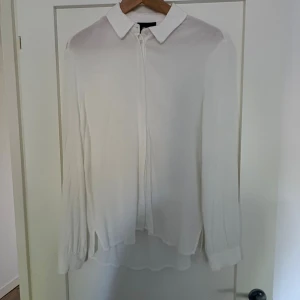 Vit skjorta med knappar och krage - Säljer en stilren vit skjorta med klassisk krage och dolda knappar framtill. Skjortan har långa ärmar med manschett och är tillverkad i ett tunt, flowy material som ger en snygg siluett. Perfekt att styla med jeans eller kjol för en clean look.