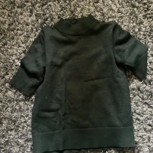 Mörkgrön polotröja från VILA - Mörkgrön polotröja från VILA med ribbade muddar vid ärmslut och nederkant. Tröjan har halvlånga ärmar och hög krage, vilket ger en stilren och enkel look. Perfekt att matcha med jeans eller kjol för en snygg outfit.