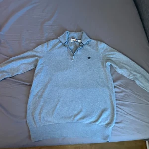 Grå half zip-tröja från Timberland - Ljusgrå långärmad tröja från Timberland i 100% ekologisk bomull. Klassisk modell med half zip och liten broderad logga på bröstet. Perfekt för lager-på-lager och har ribbade muddar vid ärmslut och nederkant.