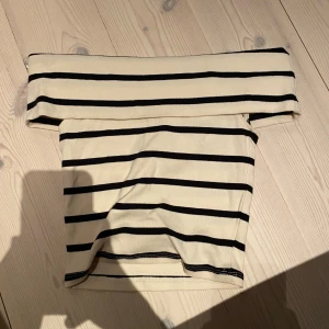 Randig offshoulder topp i beige och svart - Snygg offshoulder topp i beige med svarta horisontella ränder. Toppen är ärmlös och har en bred kant upptill som sitter snyggt över axlarna. Perfekt för en trendig och avslappnad look.