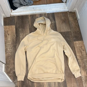 Beige hoodie med huva och ficka - Säljer en stilren beige hoodie med huva och dragsnören. Tröjan har en stor magficka framtill och är långärmad med ribbade muddar. Perfekt för chill dagar eller när du vill ha en enkel och clean look.