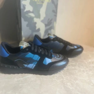 Valentino Garavani Rockrunner sneakers blå - Snygga vinter Valentino Garavani Rockrunner sneakers i svart och blått med coolt camouflagemönster. Skorna har svarta snören, detaljer i skinn och mocka samt en robust sula med nitar. Perfekta för dig som vill sticka ut med stilrena och trendiga sneakers.