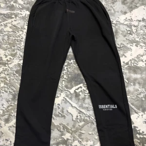 Svarta mjukisbyxor Essentials Fear of God - Svarta mjukisbyxor från Essentials Fear of God med logotyp tryckt på benet. Byxorna har elastisk midja och sidofickor, tillverkade i mjukt bomullsmaterial för maximal komfort. Perfekta för chill eller streetwear-stil.