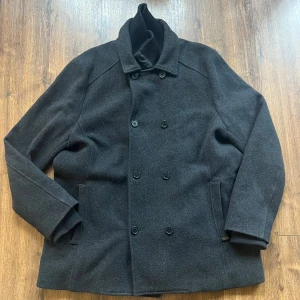 Ullrock - Stilren, tung och rejäl mörkgrå rock med dubbelknäppning och klassisk krage. Jackan har ett quiltat innerfoder med dragkedja som tyvärr är sönder och ribbade muddar vid ärmsluten. Perfekt för kyliga dagar och ger en clean look till vilken outfit som helst.