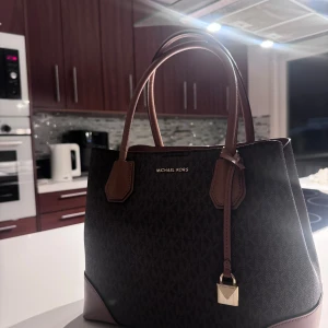 Michael Kors brun handväska med monogram - Snygg handväska från Michael Kors i mörkbrunt med klassiskt MK-monogram. Väskan har ljusbruna detaljer, dubbla handtag och guldfärgad dragkedja samt hänglås. Tillverkad i skinn och syntet, perfekt storlek för vardagens essentials.