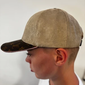 Beige keps med monogram - Snygg beige keps  med klassiskt monogrammönster i både mocka och läder. Skärmen är mörkbrun med guldiga LV-loggor och blommor, och baksidan har justerbart spänne i läder. Perfekt för dig som vill sticka ut med lyxig streetstyle.