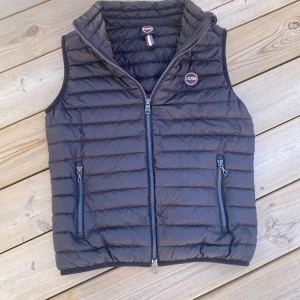 Svart dunväst från Colmar - Säljer en svart dunväst från Colmar med quiltad design och två dragkedjefickor framtill. Västen har hög krage och stängs med dragkedja. Perfekt för lager-på-lager och snygg till streetwear. Märkeslogga på bröstet.Den är i st 152. Jag köpte den på NK, har haft den 2 gånger så den är som ny. Skriv om ni undrar något