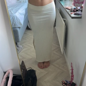 Lång ribbad kjol i ljusbeige - Säljer en lång ribbad kjol i ljusbeige med hög midja och slits på sidan. Kjolen är figurnära och har en snygg stretchig passform som framhäver siluetten. Perfekt för dig som gillar stilrena och enkla plagg med en twist.