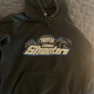 Trapstar tracksuit London Shooters - Svart hoodie från Trapstar med fet broderad London Shooters-logga i grått, blått och vitt på bröstet. Klassisk känguruficka, snörning i huvan och mjukt material. Perfekt för dig som vill sticka ut med streetwear-vibe.