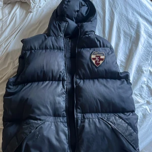 Svart dunväst från Kingsland - Säljer en svart dunväst från Kingsland med huva och broderad logga på bröstet. Västen har dragkedja framtill och två fickor. Perfekt för lager-på-lager och riktigt skön när det är kyligt ute.