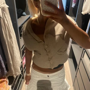 Beige croppad kofta med knappar - Säljer en beige croppad kofta med korta ärmar och stora knappar framtill. Koftan har två dekorativa fickor och en bred krage som ger en chill och trendig vibe. Perfekt att slänga över ett linne eller klänning för en snygg layering-effekt. 