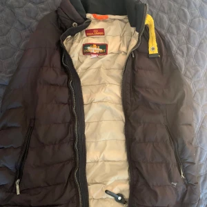 Parajumpers Väst S - Säljer en mörkbrun superlätt pufferjacka från Parajumpers i storlek S. Jackan har quiltad design, dragkedja framtill och två sidofickor med dragkedja. Insidan är ljusbeige och jackan har en gul detalj vid kragen. Perfekt för kyliga dagar!