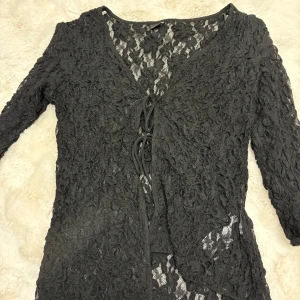 Svart spetsblus från Gina Tricot - Säljer en svart blus från Gina Tricot i storlek M. Blusen är helt i spets med blommigt mönster, har v-ringning och snörning framtill. Långärmad och transparent, använts fåtal gånger.