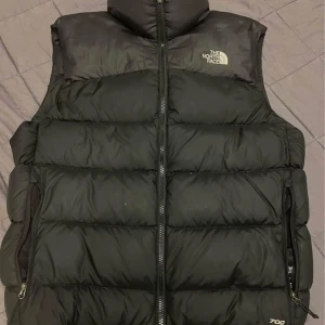 Svart dunväst från The North Face - Säljer en svart dunväst från The North Face, modell 700. Västen har hög krage, två dragkedjefickor och tydlig logga på bröstet. Klassisk quiltad design med breda kanaler och hel dragkedja framtill. Perfekt för lager-på-lager under kyliga dagar.