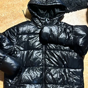 Svart pufferjacka med huva - Säljer en svart pufferjacka med glansig finish och huva. Jackan har dragkedja framtill och breda, quiltade sektioner som ger en riktigt fet och puffig look. Perfekt för kalla dagar när du vill hålla värmen och ändå se cool ut.